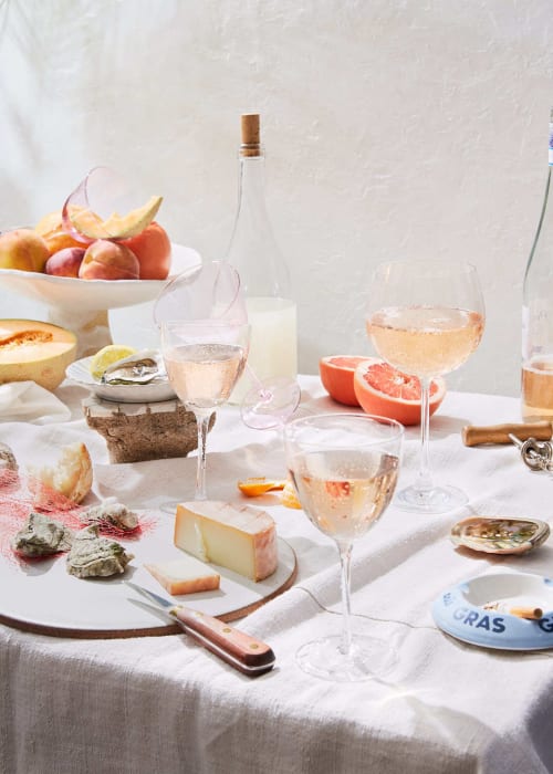 Still life with various cheeses, oysters, wine glasses, and assorted fruits such as peaches and grapefruits on a white tablecloth.
