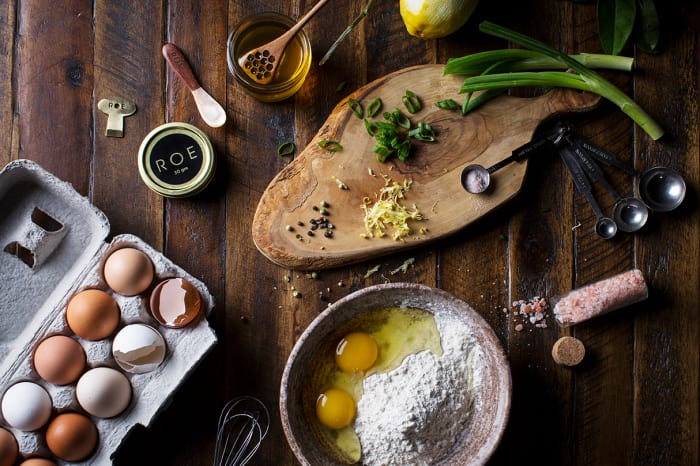 Overhead shot of baking ingredients: eggs in carton, flour, honey, lemon, scallions, and spices arranged on a rustic wood surface.
