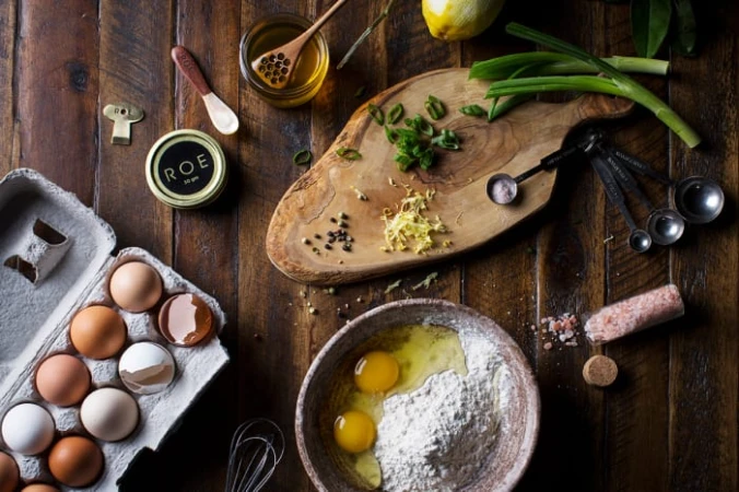 Overhead shot of baking ingredients: eggs in carton, flour, honey, lemon, scallions, and spices arranged on a rustic wood surface.