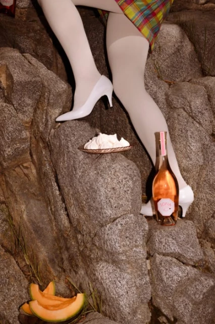 Fashionable scene on rocks featuring a model's legs, white heels, cantaloupe, wine bottle, and a plate of meringue desserts.