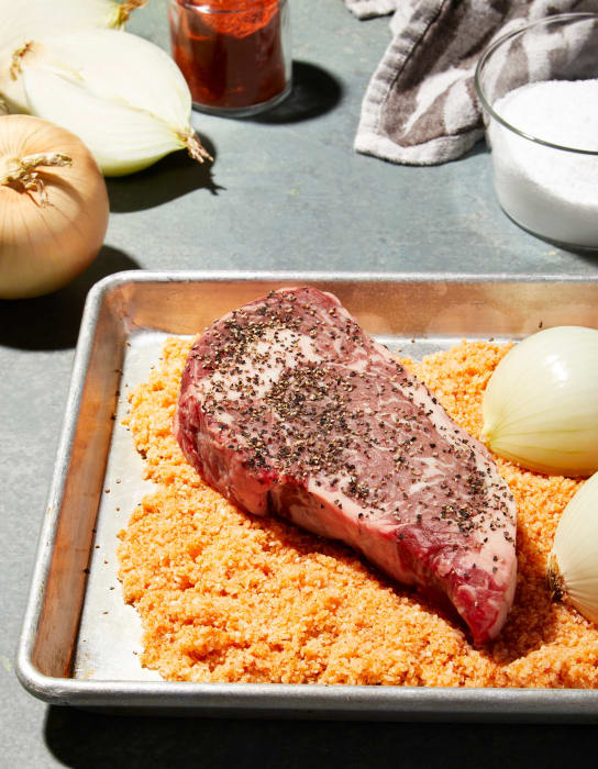 Raw steak sits on a metal tray with orange seasoning and onions, surrounded by spices and a towel on a grey surface.