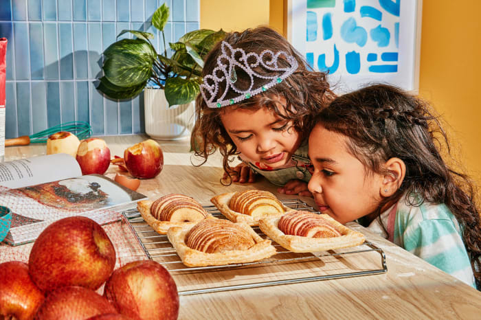 Two young girls in a kitchen look at apple pastries on a cooling rack, with apples, cooking book, and kitchenware visible.
