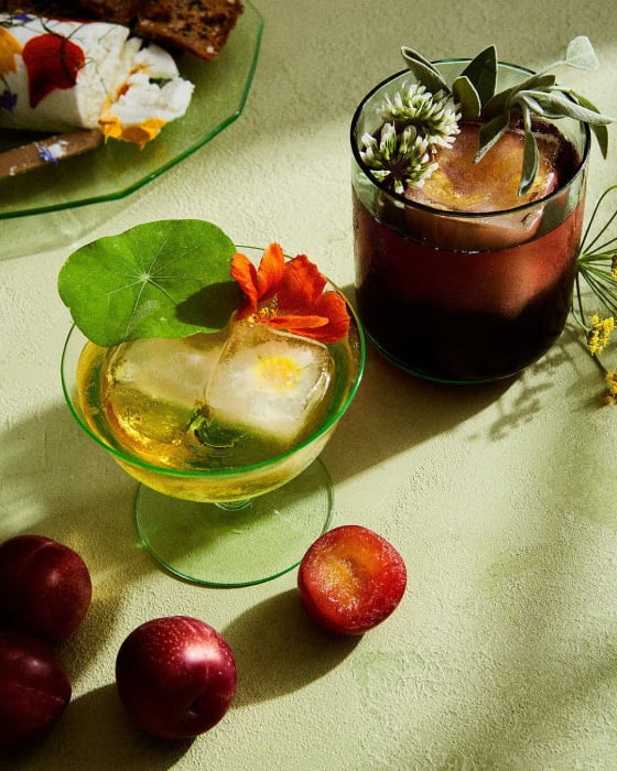 Two cocktails, one yellow and one red, sit on a green surface with plums and a cheese log on a plate, both garnished with flowers.