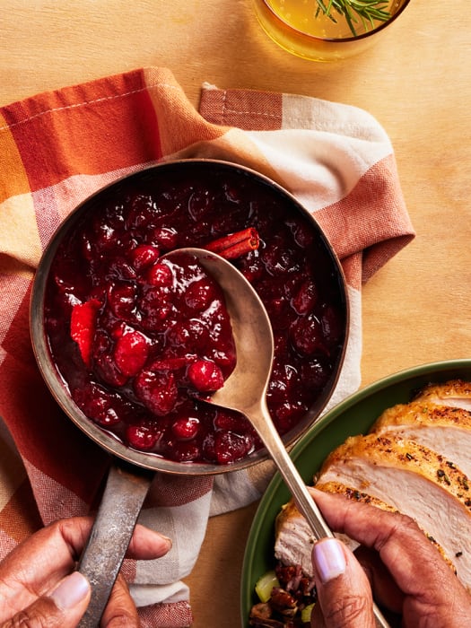 Overhead shot of cranberry sauce being served with a spoon next to sliced turkey on a table with a patterned napkin and juice.