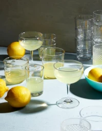 Several clear glassware glasses filled with yellow cocktail and whole yellow lemons scattered on a table under direct light