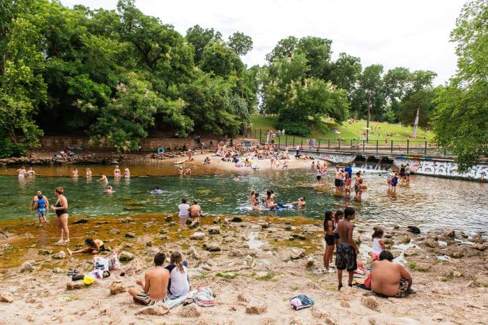 A large group of people of varying ages swim and relax in a natural spring surrounded by a sandy bank and lush green trees.