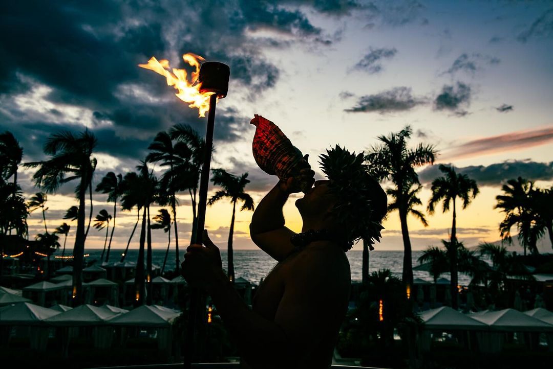 Silhouette of a Hawaiian man blowing a conch shell and holding a flaming torch at sunset, with palm trees and ocean in the background.