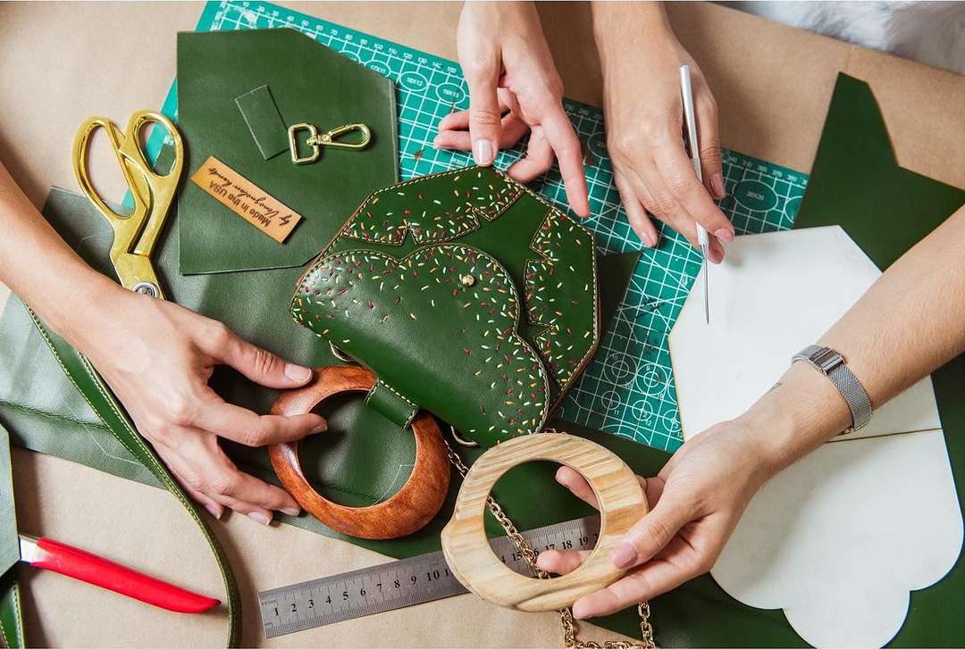 Two people are crafting a green leather purse indoors, using a pattern, ruler, and scissors, with wood purse handles on a craft mat.
