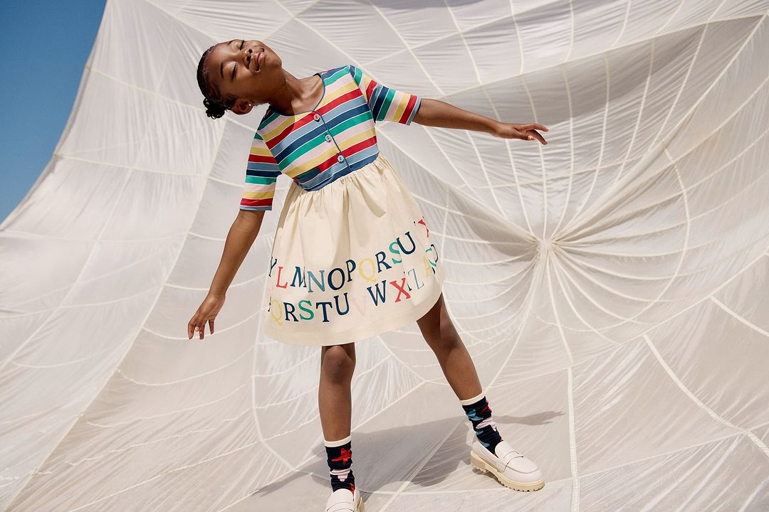 Young girl in striped shirt and alphabet skirt poses in front of a parachute, showcasing children's fashion against a white backdrop.