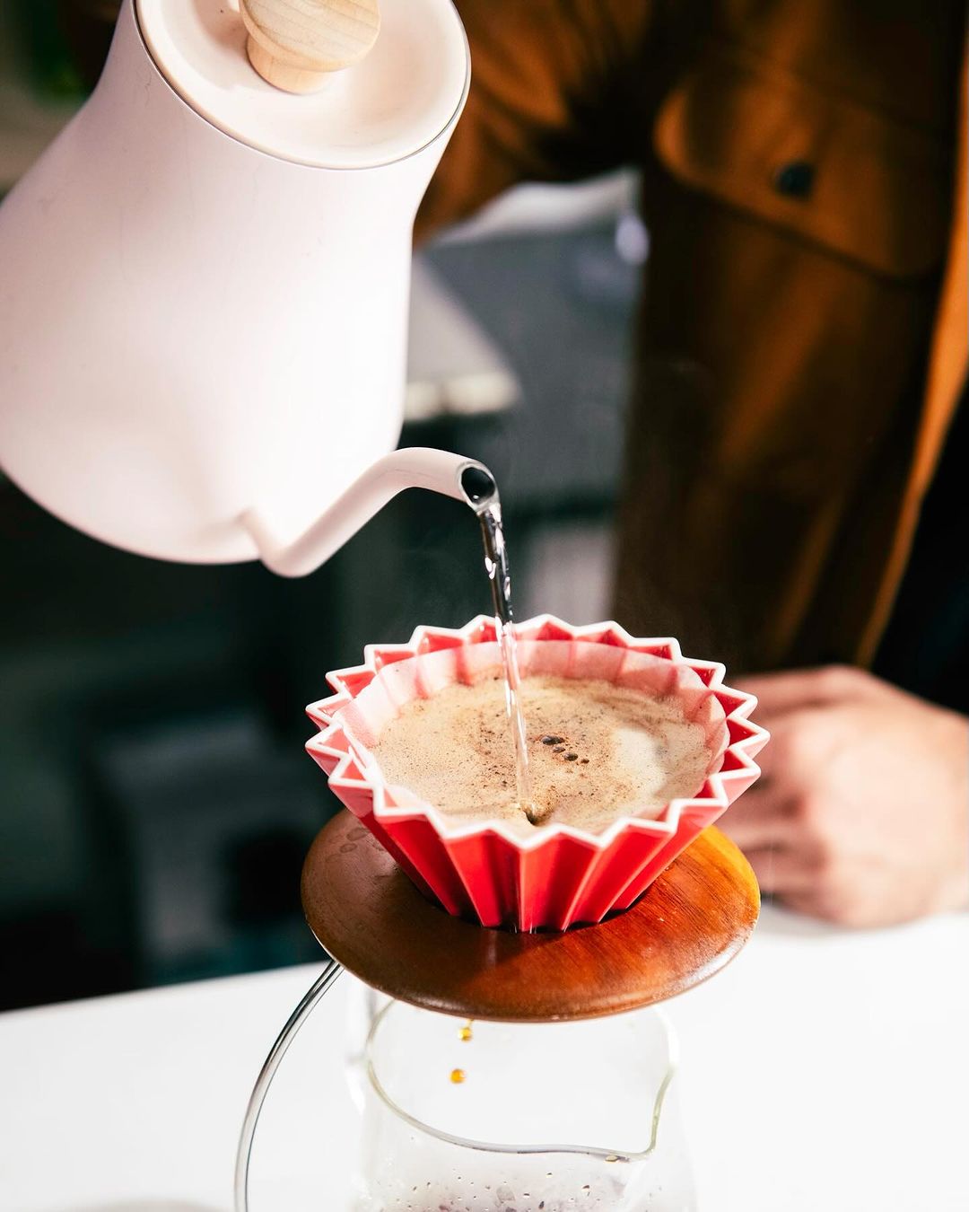 Water is poured from a white kettle into a red origami paper filter, brewing coffee in a glass server on a white counter.