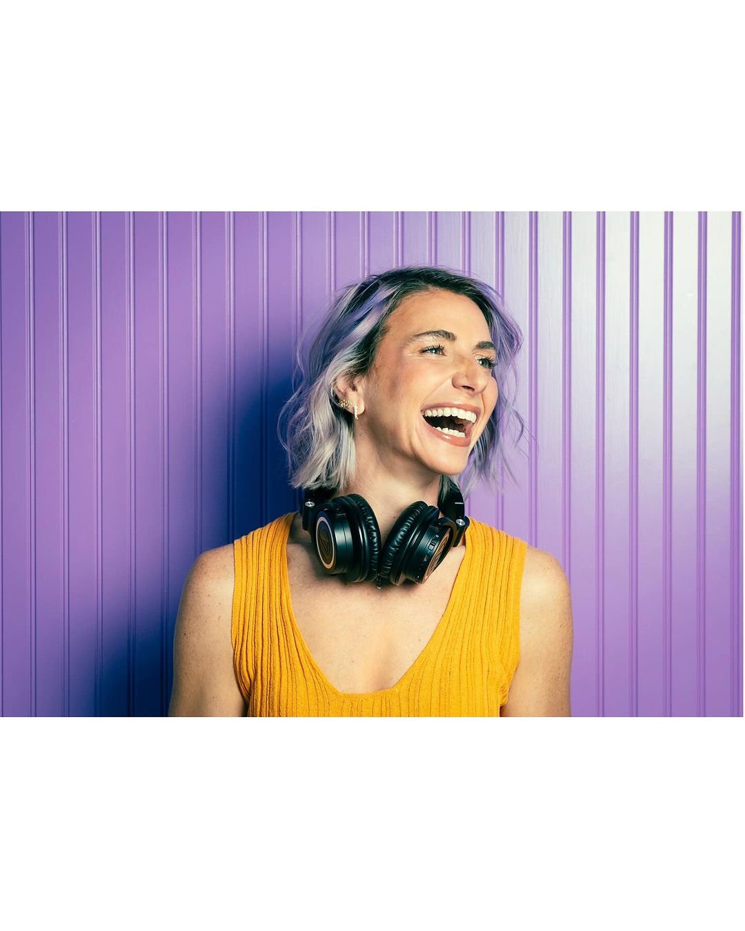 Laughing woman with headphones and purple hair stands against a purple paneled wall in a medium shot, looking off camera.