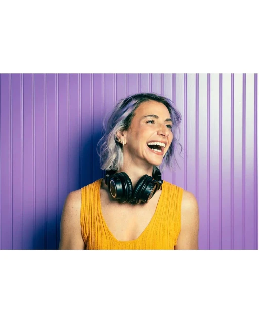 Laughing woman with headphones and purple hair stands against a purple paneled wall in a medium shot, looking off camera.