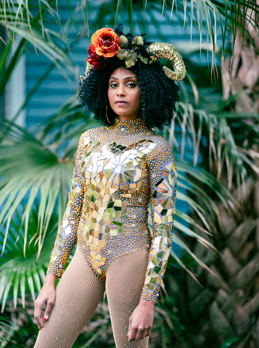 Woman in a gold sequin bodysuit with floral headdress and goat horns, standing in front of palm trees in an outdoor setting.