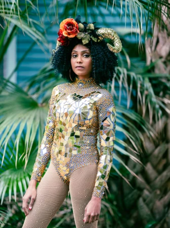 Woman in a gold sequin bodysuit with floral headdress and goat horns, standing in front of palm trees in an outdoor setting.