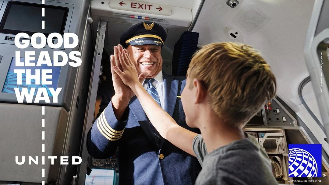 Airline pilot smiles and gives a high-five to a child in the aisle of an airplane; travel and family advertisement with airline branding.