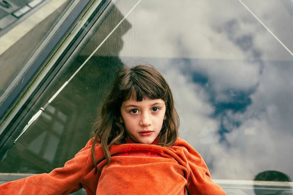 Portrait of a young girl with brown hair and bangs, wearing an orange sweatshirt, sitting indoors with a reflective window and city view.
