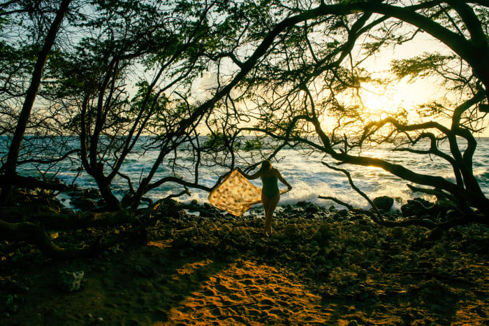 Woman in swimsuit standing on rocky beach with patterned wrap, framed by silhouetted trees against a bright sky and ocean waves.