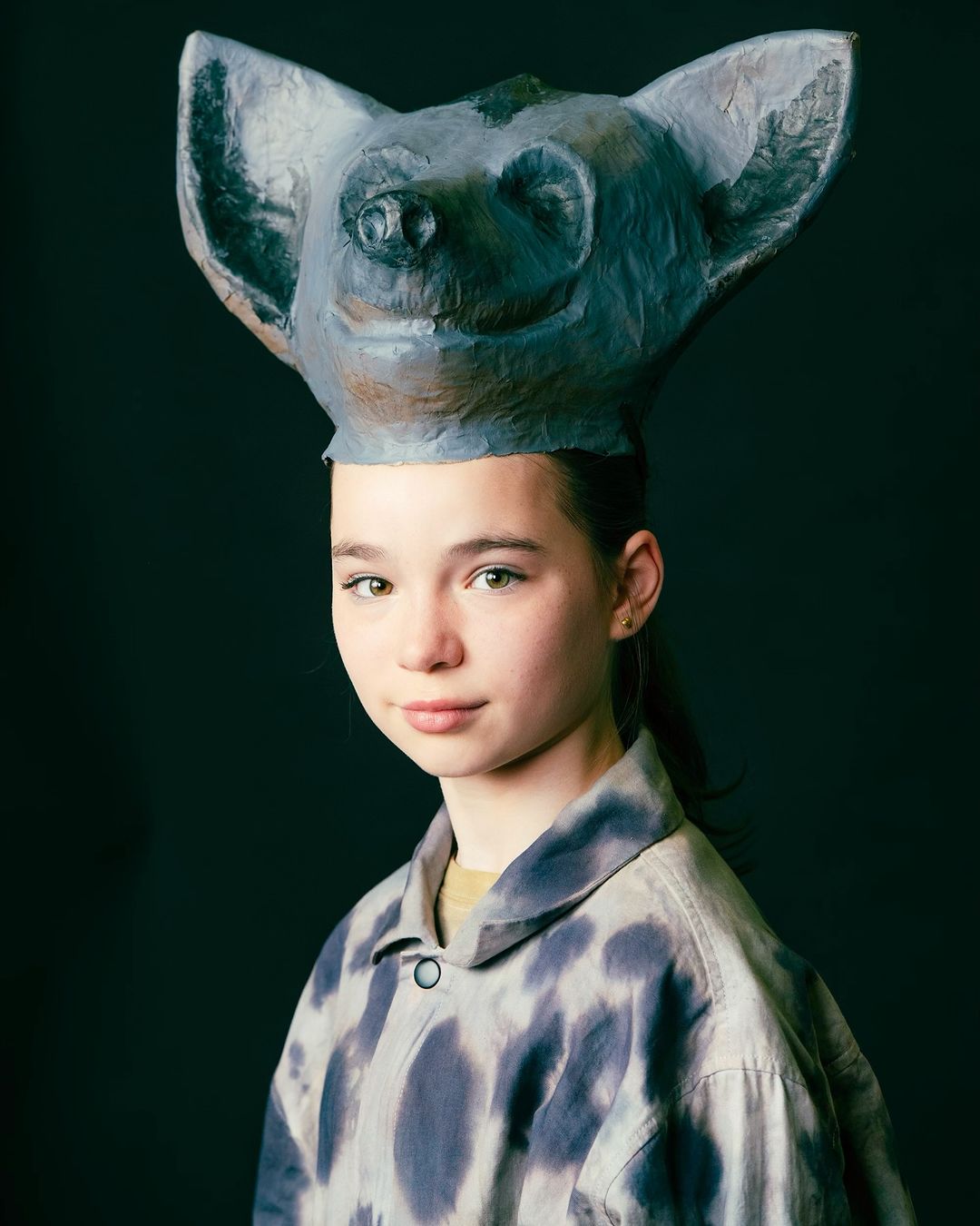 Young woman wearing an oversized tie-dye jacket and a large paper mâché mask in the shape of a cartoonish creature against a dark backdrop.