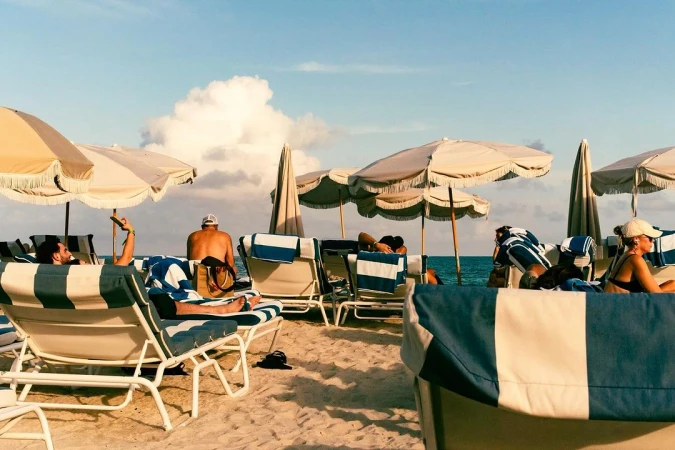Several people lounge on striped beach chairs under umbrellas on a sunny day at the beach, with the ocean and sky in the background.