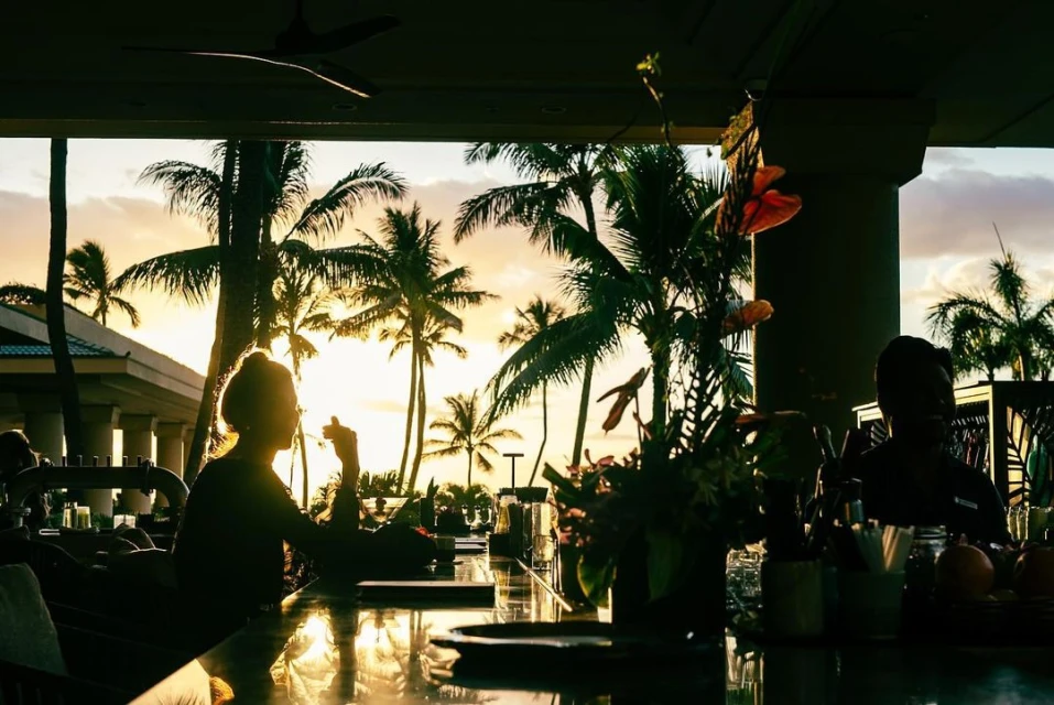Silhouetted figures at an open-air bar at sunset with palm trees, tropical plants, and island architecture in the background.
