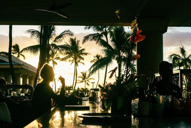 Silhouetted figures at an open-air bar at sunset with palm trees, tropical plants, and island architecture in the background.