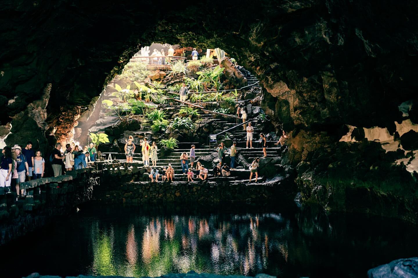 Scenic view inside volcanic cave with group of people on steps, lush tropical plants, and pool reflecting sunlight in tranquil setting.