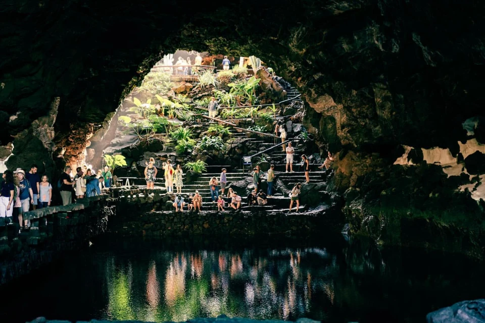 Scenic view inside volcanic cave with group of people on steps, lush tropical plants, and pool reflecting sunlight in tranquil setting.