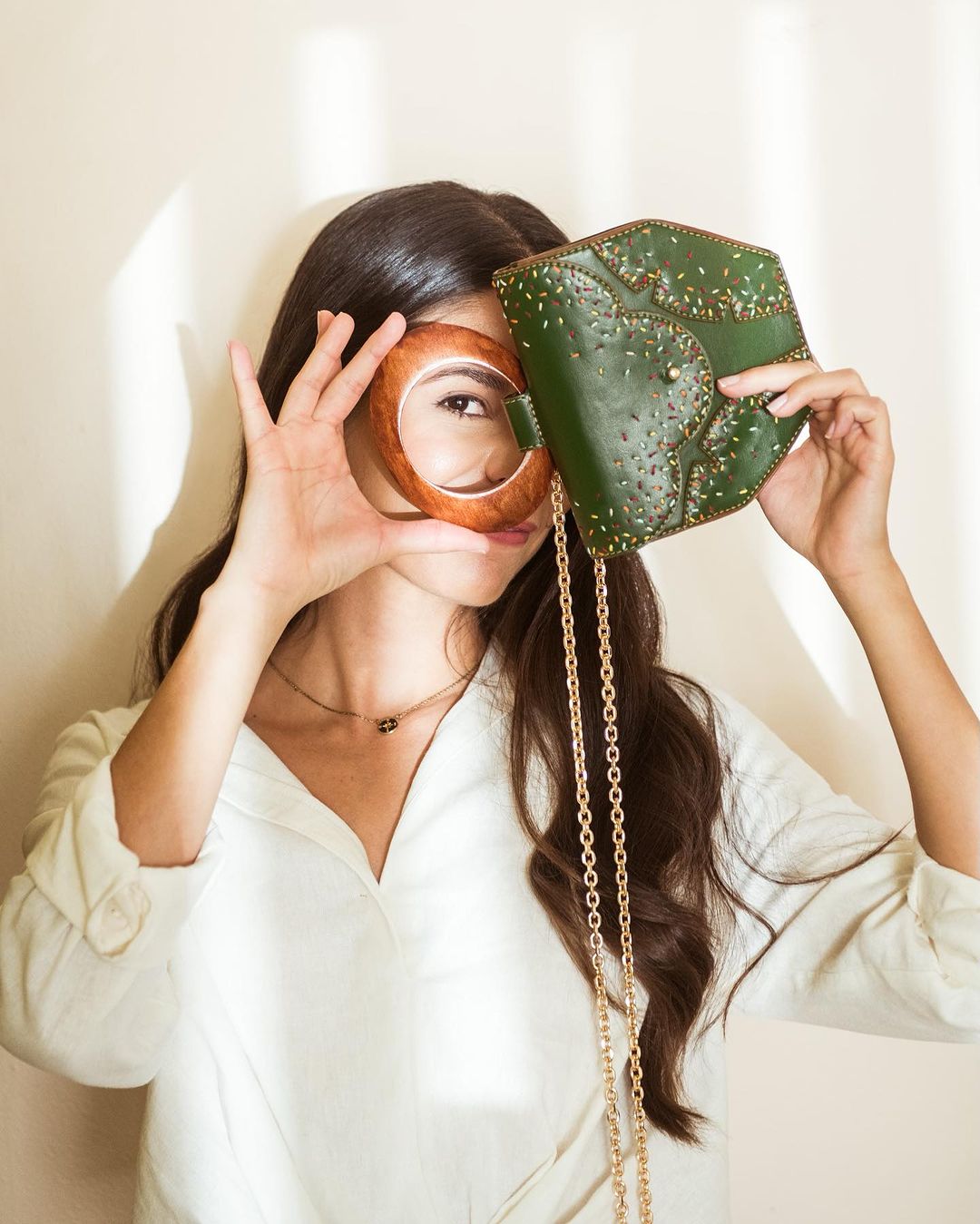 A young woman with dark hair holds a green handbag and a wooden bangle in front of a cream wall indoors with natural lighting.
