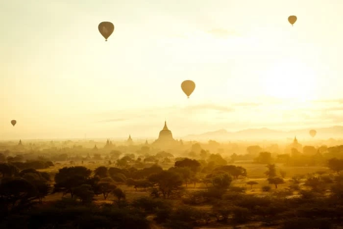 Scenic aerial landscape featuring hot air balloons at sunrise with ancient temples and lush trees visible below a hazy sky.