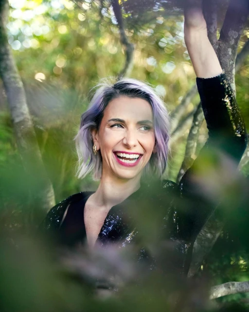 Smiling woman with purple hair poses in nature, wearing sequin top. Lush green leaves frame the shot, creating a bokeh background.