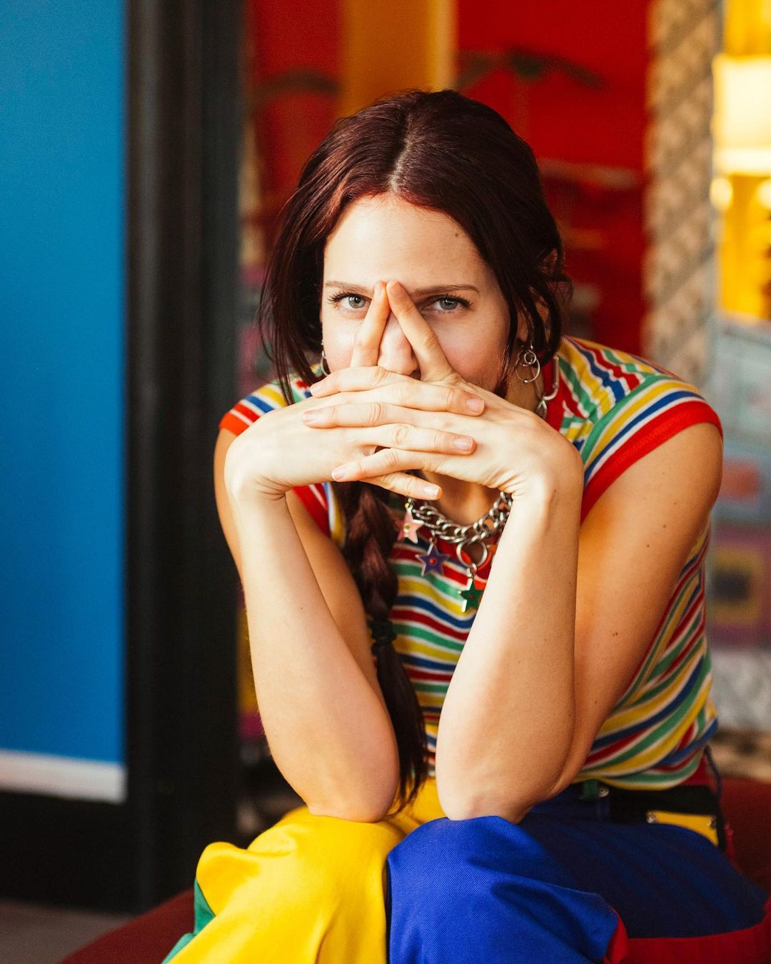 Portrait of a woman with auburn hair, wearing a colorful striped shirt and a chain necklace, sitting indoors with her hands raised.