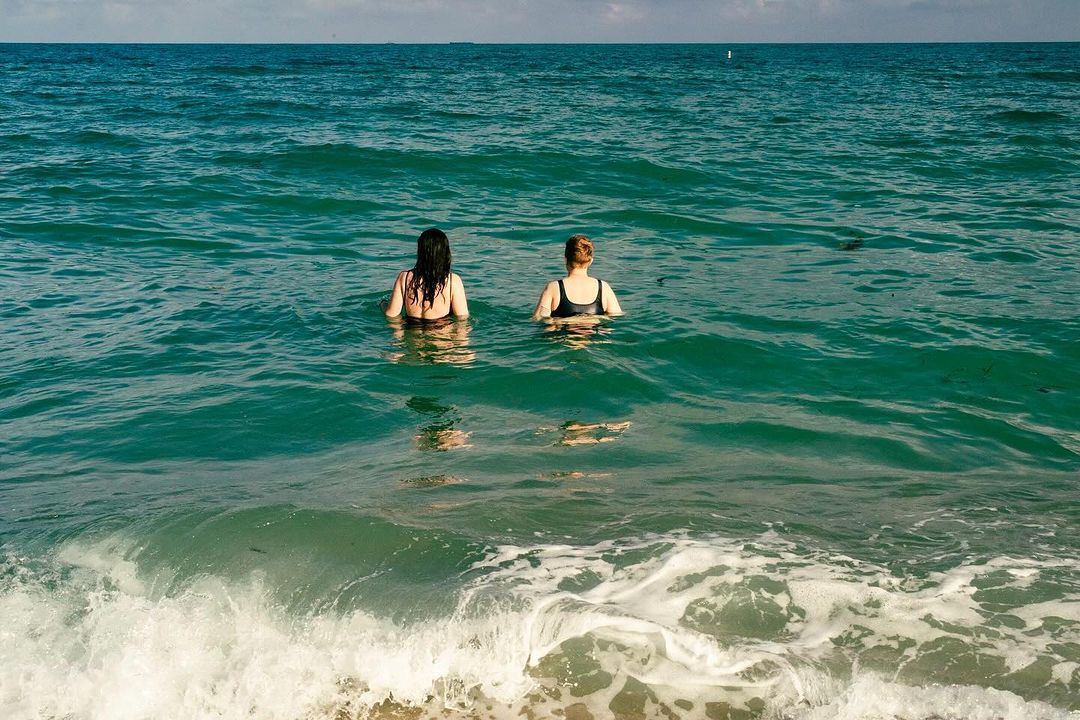 Two women swimming in turquoise ocean water with waves breaking in the foreground captured by Gabriela Herman
