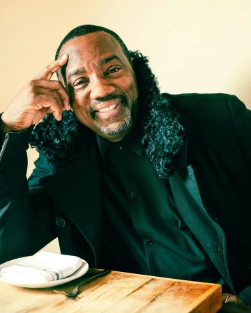 Portrait of a smiling Black man in a black jacket seated at a wooden table with a plate and fork.