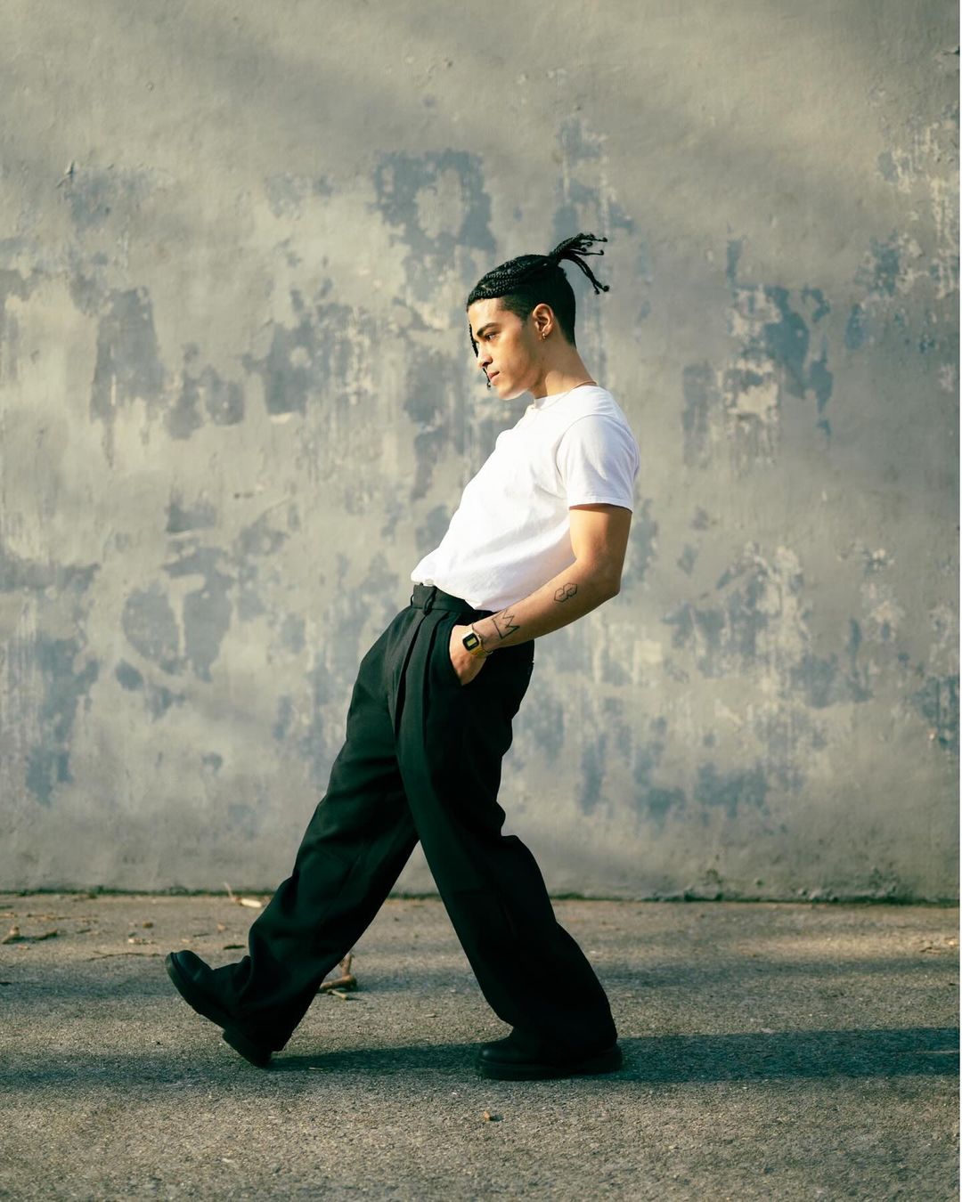 A young adult man with braided hair poses in a white t-shirt and black pants, set against a weathered wall, with an urban street surface.