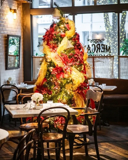 Christmas tree in a restaurant setting, adorned with red and gold decorations, standing near tables and chairs, creating a festive ambiance.