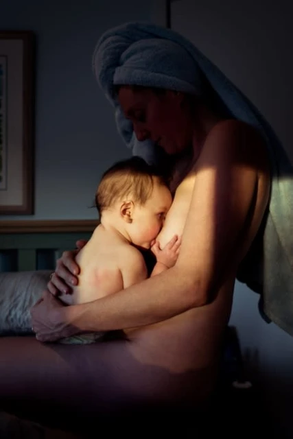 Woman with a towel on her head breastfeeds a baby indoors in a dimly lit room with sunlight on her skin, capturing a tender moment.