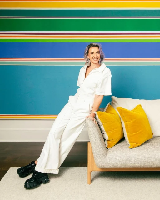 Smiling woman in white jumpsuit leans on a neutral couch in a colorful, modern interior setting with abstract striped wall art.