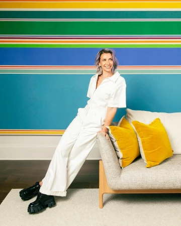 Smiling woman in white jumpsuit leans on a neutral couch in a colorful, modern interior setting with abstract striped wall art.