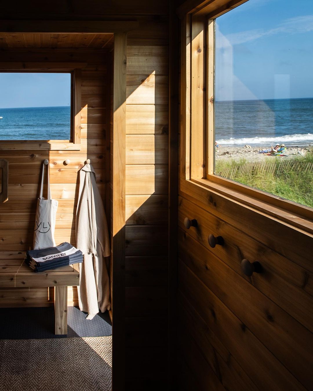 Wooden interior with a beach view through windows, bench with folded towels and tote bag, and light streaming through the windows.