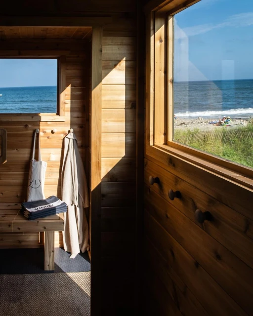 Wooden interior with a beach view through windows, bench with folded towels and tote bag, and light streaming through the windows.