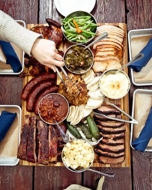 Overhead shot of a wooden board laden with barbecue meats like ribs, sausages, brisket, pulled pork, and sides like mac and cheese.