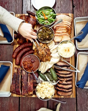 Overhead shot of a wooden board laden with barbecue meats like ribs, sausages, brisket, pulled pork, and sides like mac and cheese.