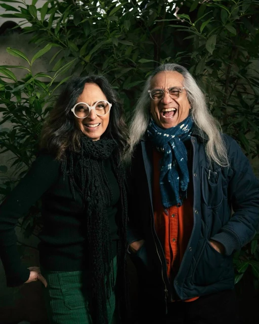 Portrait of a smiling couple in front of green plants, woman with glasses and black scarf, man with long gray hair and blue scarf.