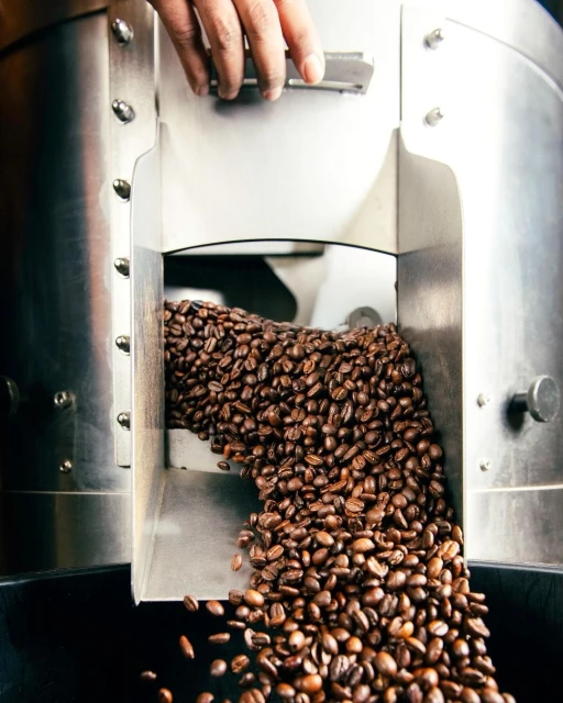 Close-up of roasted coffee beans spilling from a stainless steel industrial roaster, highlighting texture and color in a dark, industrial setting.