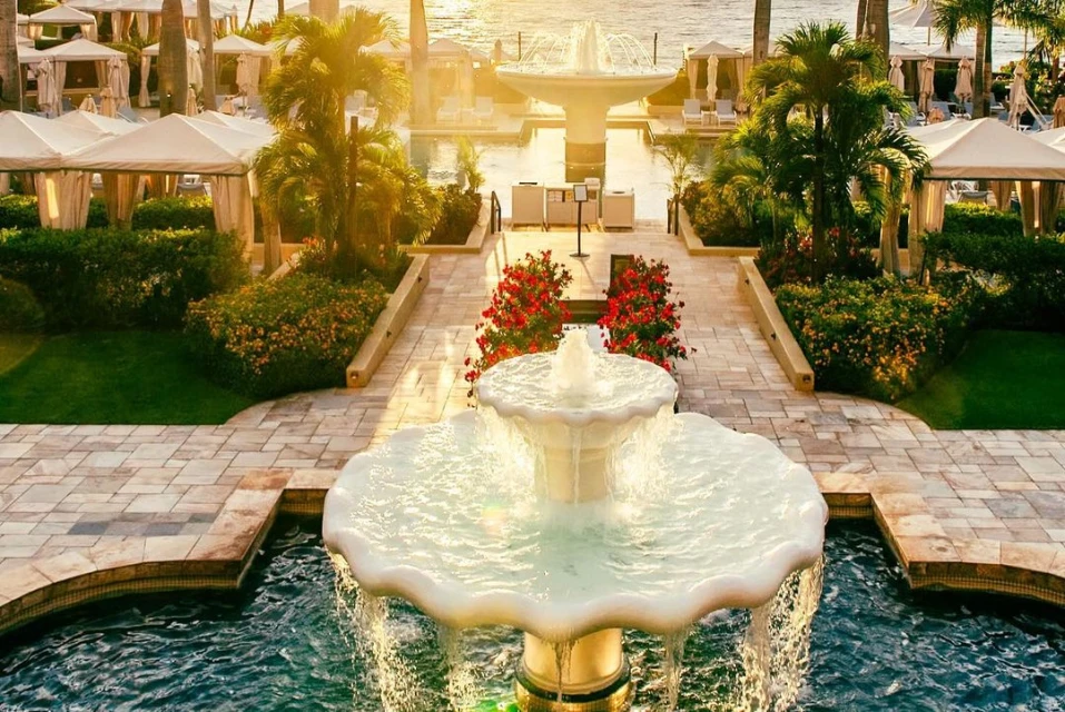 Elegant outdoor fountain at a luxury resort with manicured gardens, walkways, and poolside cabanas, with the ocean visible in the background.