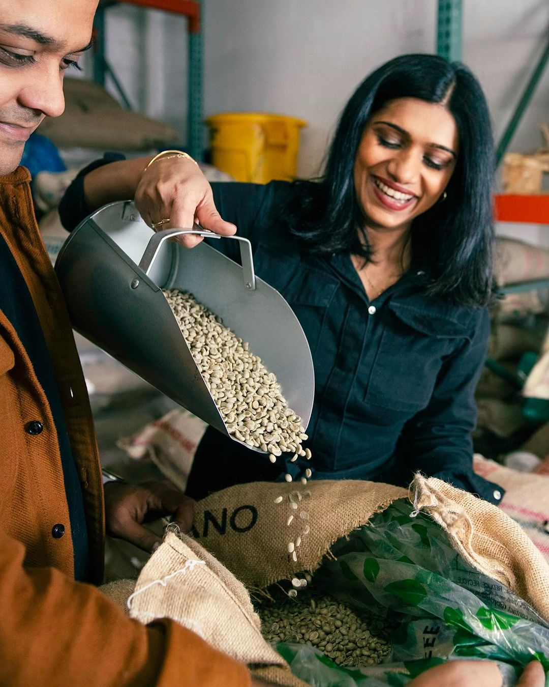 Two adults smiling while pouring coffee beans from a metal scoop into a burlap sack inside a warehouse with shelving in background.