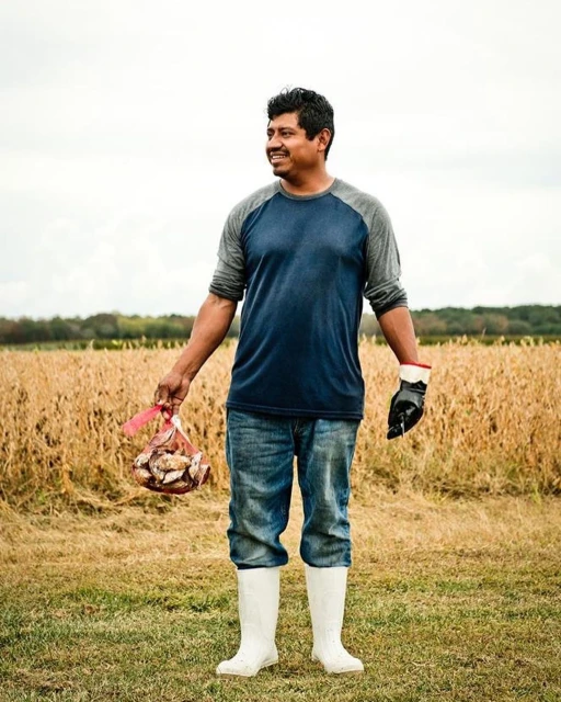 Full shot of a smiling Hispanic man wearing white boots, jeans, and a raglan shirt, holding a bag of mushrooms, posing outdoors.