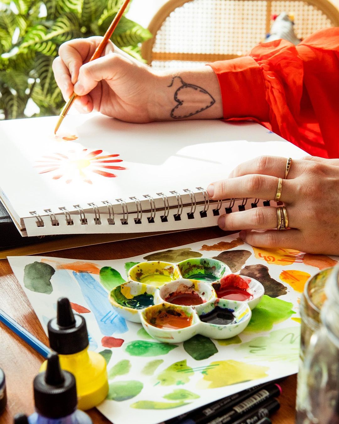 Person painting a vibrant sun on paper with watercolors in an indoor space with art supplies and bright natural light, red shirt.
