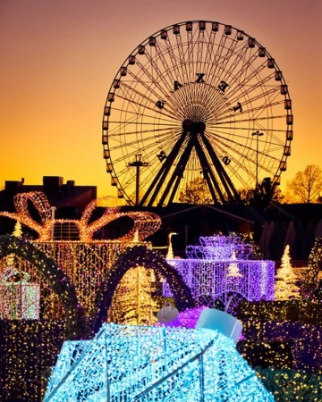 Illuminated ferris wheel stands against a vivid sunset sky, with glowing gift box decorations in the foreground at night.