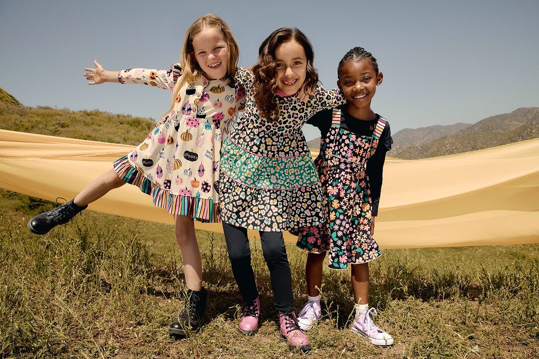 Three cheerful girls in floral dresses pose outdoors in a grassy field on a sunny day, wearing colorful patterned outfits with mountains visible.
